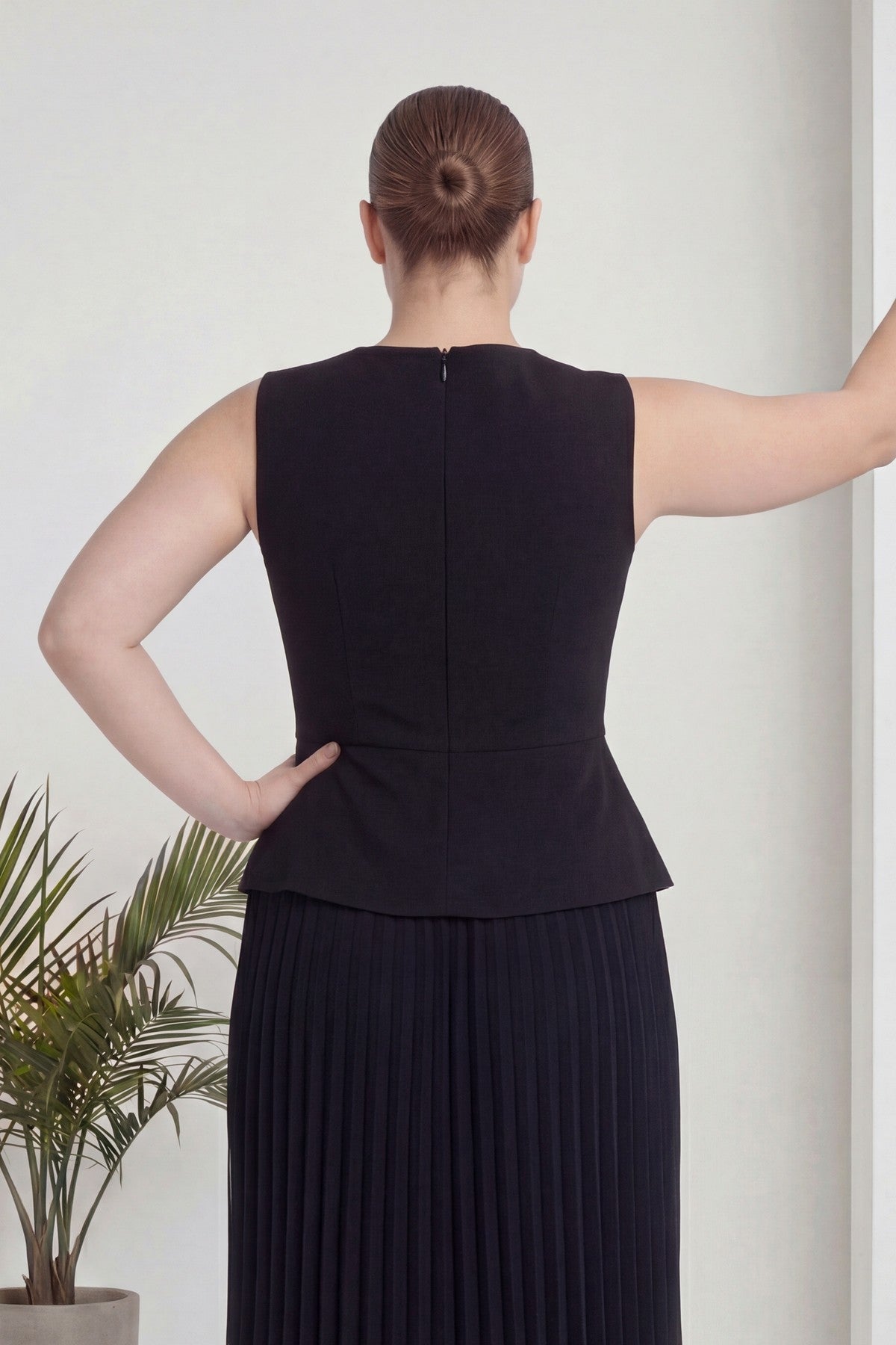 Siyah büyük beden piliseli kolsuz midi elbise ar, topuz saçlı, bir eli belinde çiçek önü.