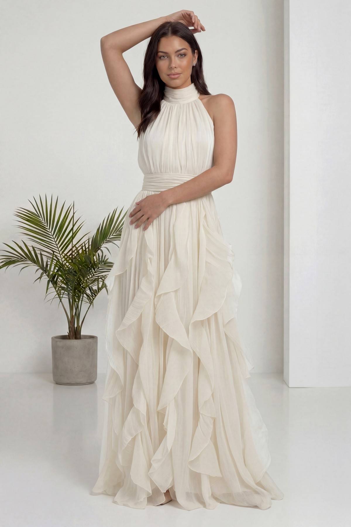 The ZKS ekru halter yaka fırfırlı uzun abiye elbise, sol eli belde, sağ kol havada poz