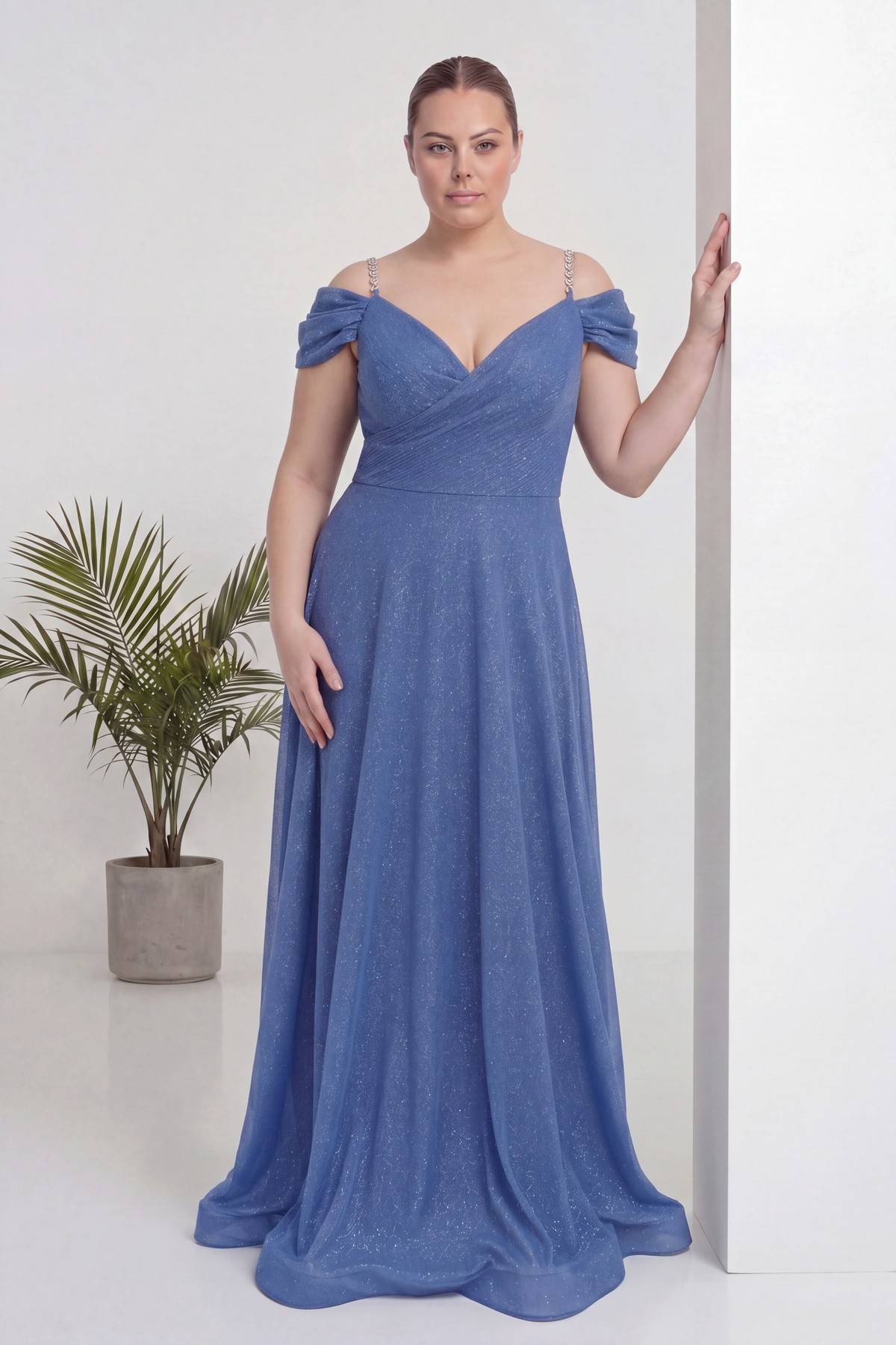 The ZKS indigo büyük beden kloş piliseli kruvaze abiye elbise, omuzları açık, V yaka, model duvara yaslanmış