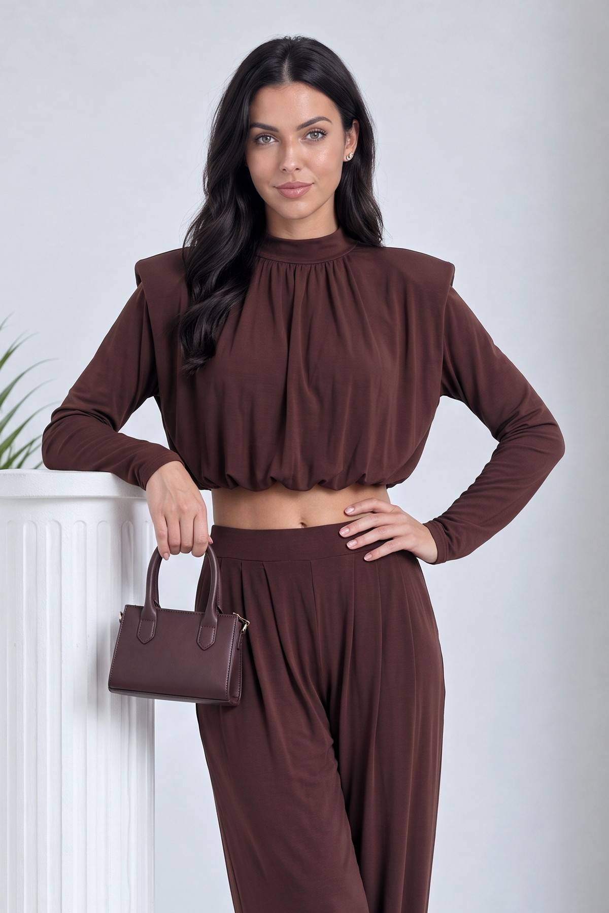 Kadın kahverengi crop şalvar takım, büzgülü yüksek yaka detaylı, model elinde küçük çanta tutuyor.