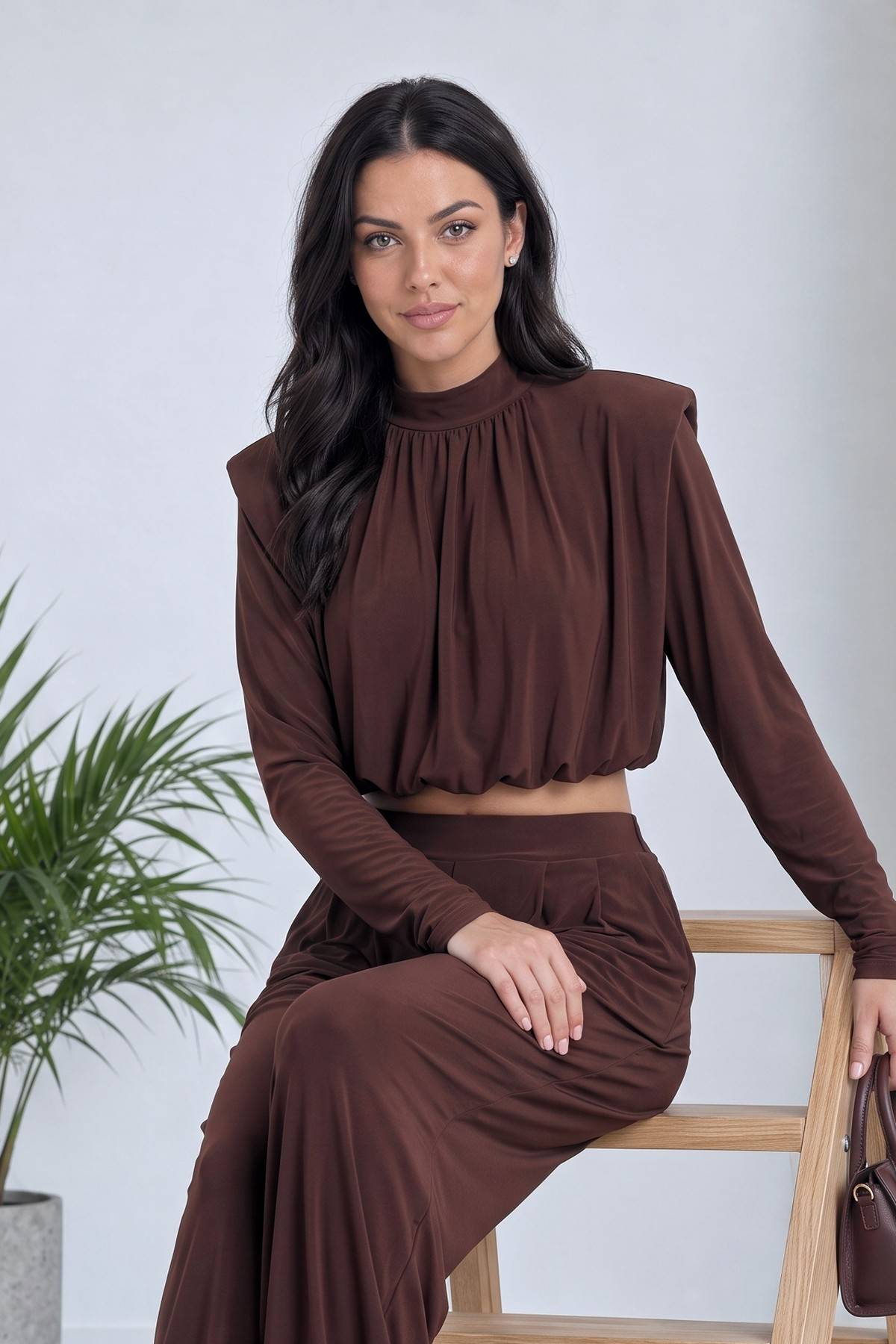 Model kahverengi crop şalvar takımıyla ahşap sandalyeye oturmuş, elinde çanta var.