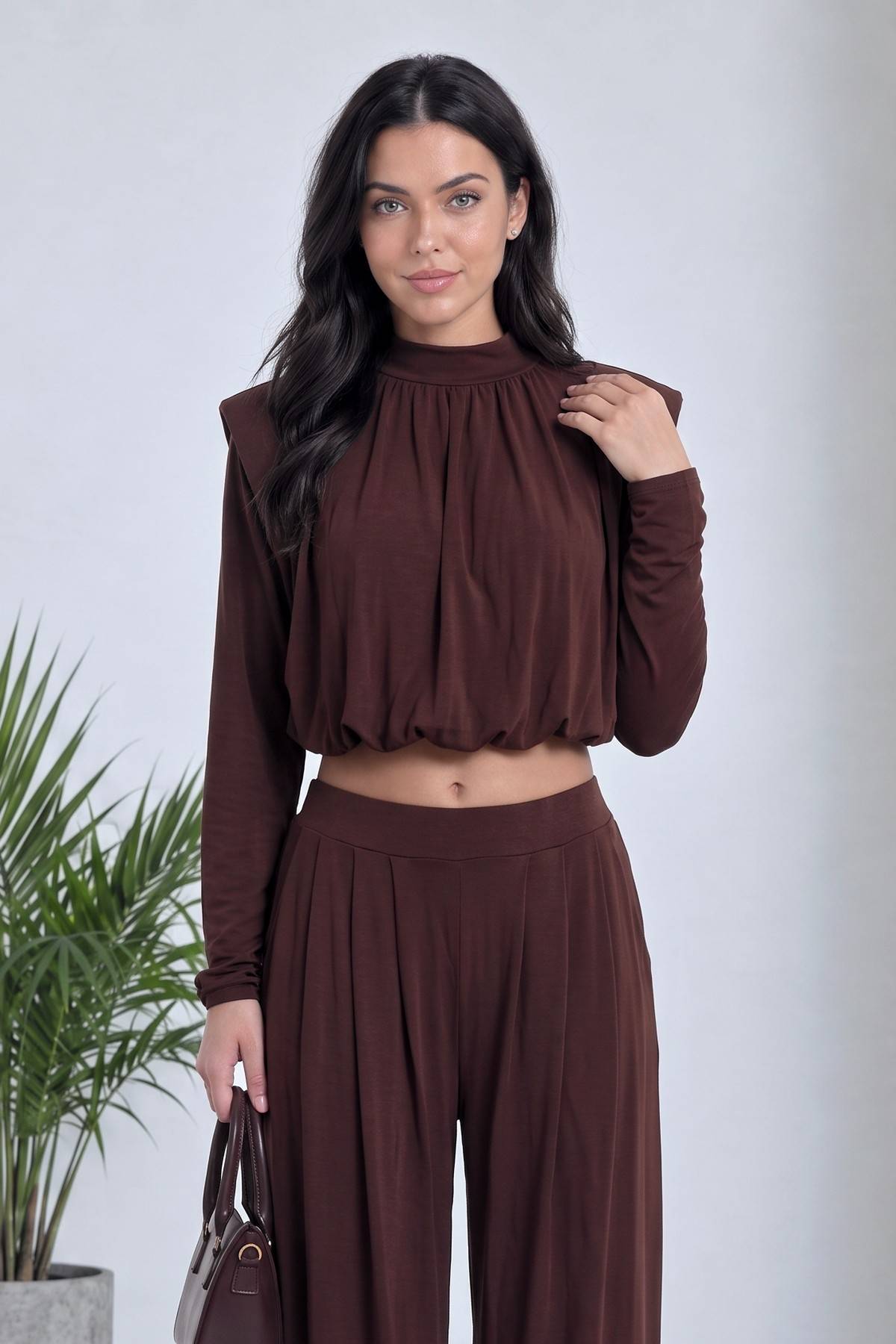 Kadın kahverengi crop şalvar takım, büzgülü yüksek yaka detaylı, model elinde küçük çanta tutuyor, yandan hafif açılı.