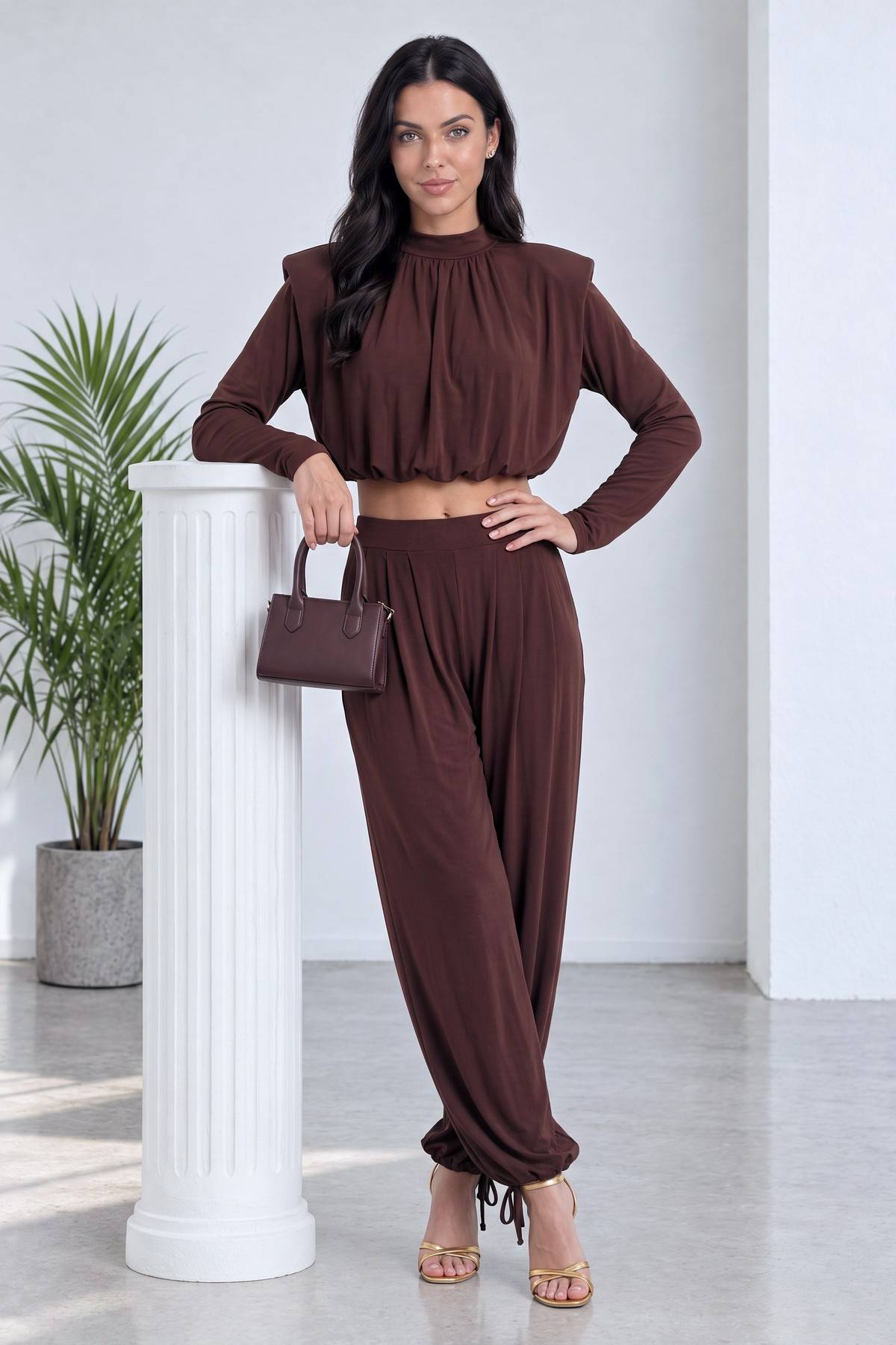The ZKS kahverengi crop şalvar takım, omuz vatkalı, büzgülü yakalı, model elinde çanta ile ayakta duruyor.