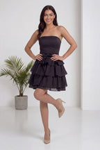 Model, The ZKS straplez siyah fırfır etekli kısa abiye elbiseyi önden giymiş, saçını düzeltirken elbiseye odaklanıyor.